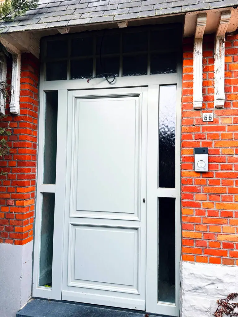 Lichtgrijze houten voordeur met zijlichten en bovenlicht in een bakstenen gevel